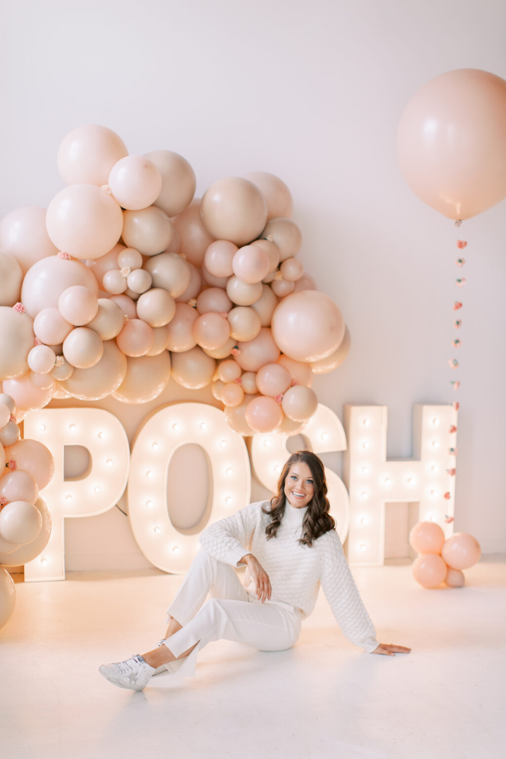 The Posh Balloon owner sitting beside a glowing POSH marquee and blush balloon installation
