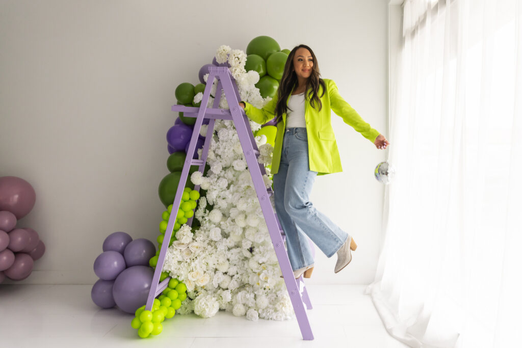 Subtle Flex Designs owner standing beside a lavender ladder styled with white florals and green and purple balloons