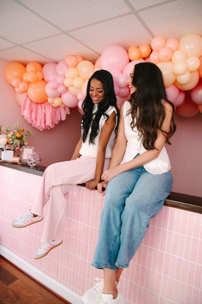 Pop Clink Events owners sitting on their pink tiled bar in front of a peach balloon garland and tassel tails